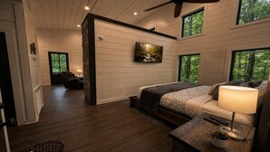A bedroom with a bed, a bedside table with a lamp, a painting on the wall, and a view of the outdoors through a window. - Romantic Cabin with Hot Tub, Fire Pit, BBQ & Fenced Yard (Broken Bow)