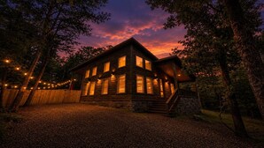 A wooden cabin with multiple windows, a stone foundation, and a wooden deck. - Romantic Cabin with Hot Tub, Fire Pit, BBQ & Fenced Yard (Broken Bow)