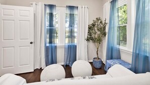 A room with a white door, two windows with blue curtains, a potted plant, and a white sofa with a patterned cover. - Peaceful Lake House - Sleeps 10, Games, Fishing & Near Downtown/Airport/BJCC (Birmingham)