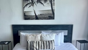 Een net opgemaakt bed met een witte deken en kussens, een geweven mand op het bed, en een schilderij van palmbomen en een strand erboven.