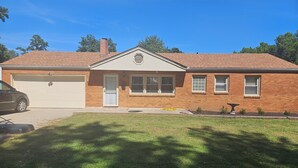 A single-story brick house with a garage, a front door, and a small front yard with a lawn and a decorative planter. - Raytown Ranch - Mahome Away from Home (Raytown)