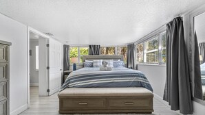 A bedroom with a large bed, a wooden headboard, a white carpet, and a view of greenery through the windows.