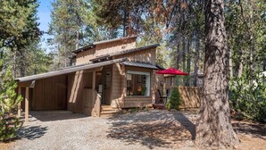 A wooden cabin with a covered porch, a red umbrella, and a gravel driveway surrounded by trees. - Steps to Village, A/C, Hot Tub, 6 SHARC, Bikes (Sunriver)