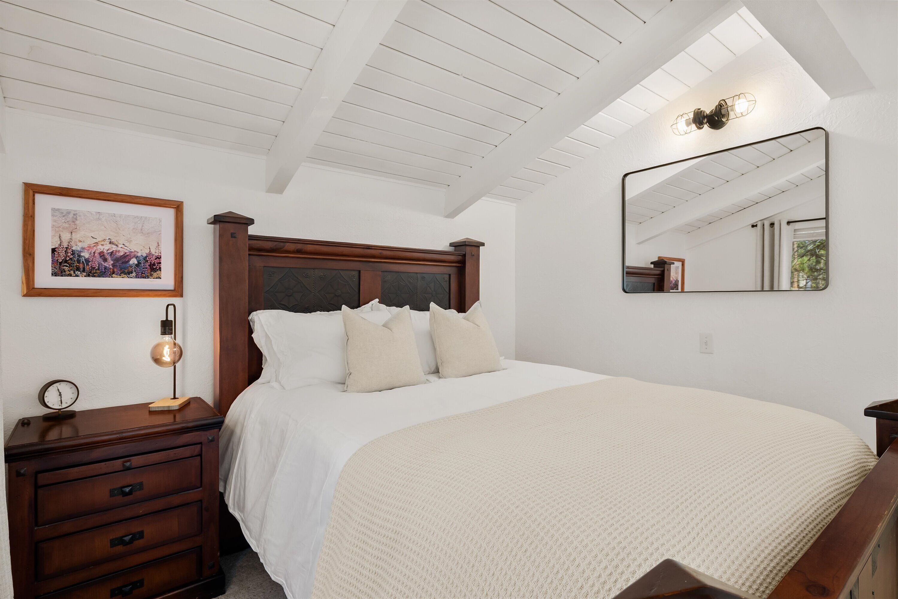 A bedroom with a wooden headboard, a bedside table with a lamp, a framed picture on the wall, and a large mirror.