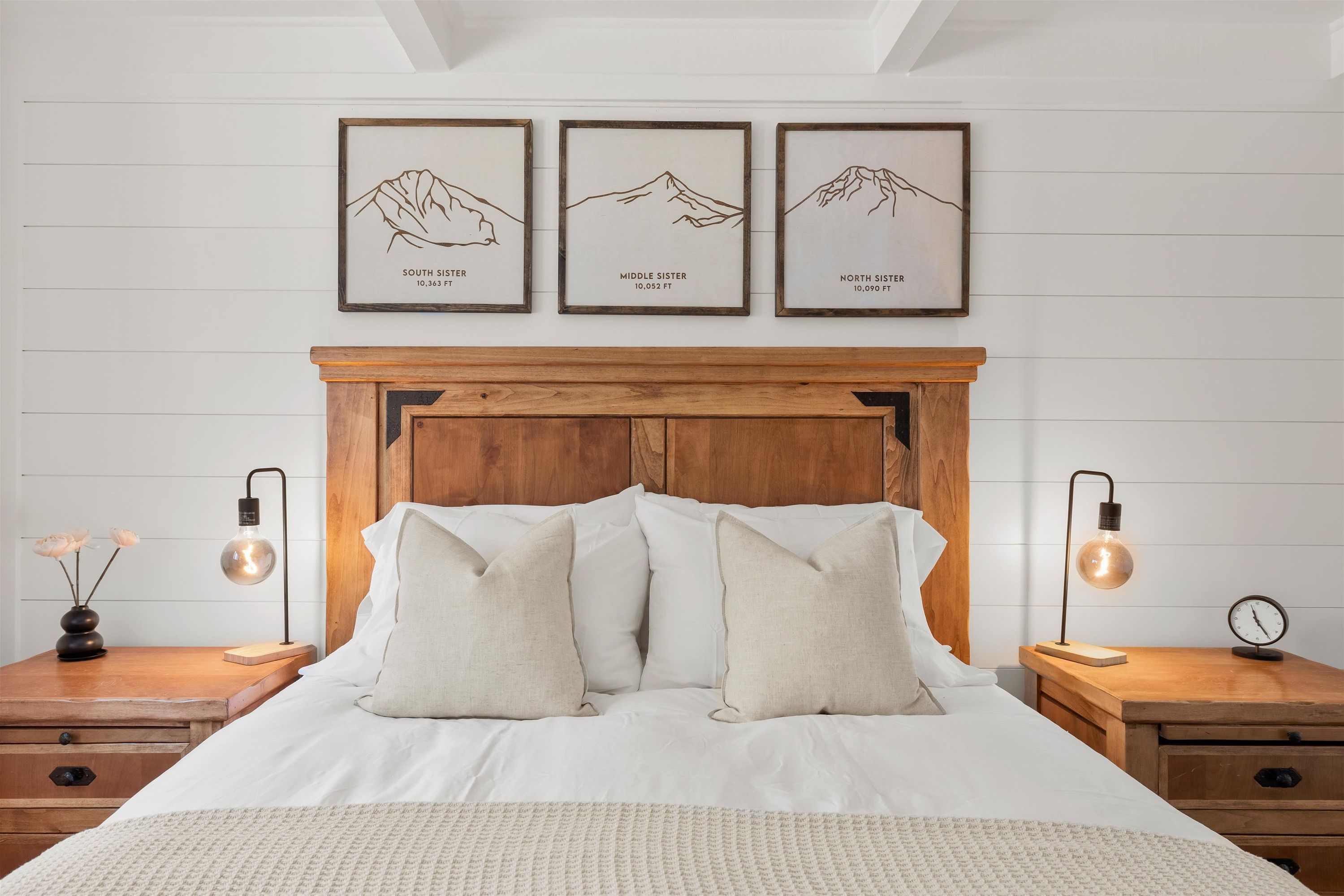 A neatly made bed with a wooden headboard, two bedside tables with lamps, and three framed pictures of mountains labeled South Sister, Middle Sister, and North Sister.