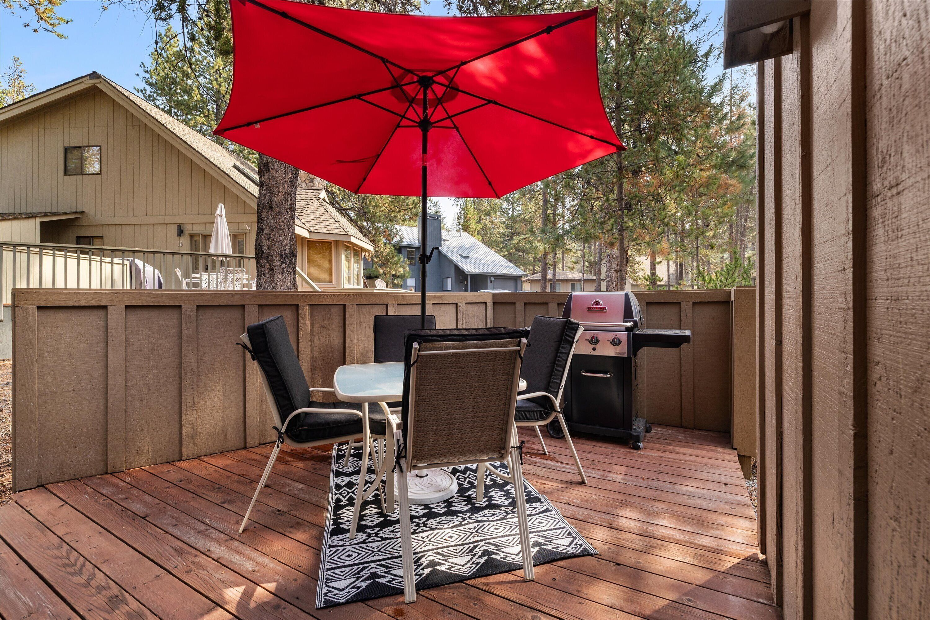 Eine Holzterrasse mit Tisch, Stühlen, einem roten Sonnenschirm und einem Grill.