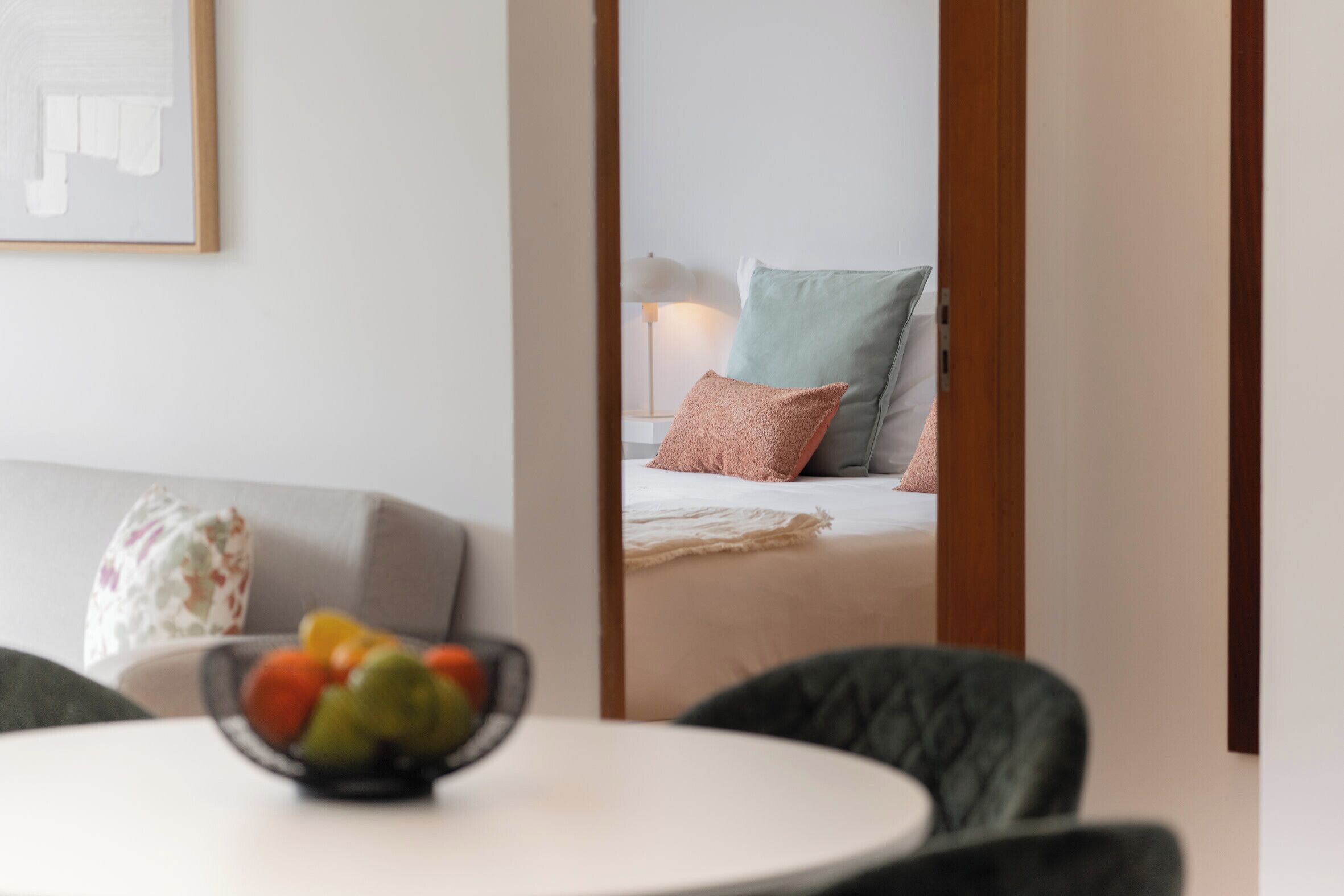 A room with a bed, a bedside table with a bowl of fruit, a lamp, and a mirror reflecting the room.