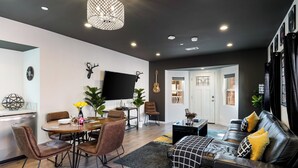Een moderne woonkamer met een zwarte leren bank, een eettafel met stoelen, een flatscreen televisie aan de wand en een kroonluchter.
