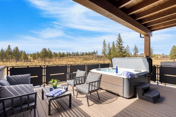 A covered outdoor deck with a hot tub, seating area, and a view of a forested landscape.