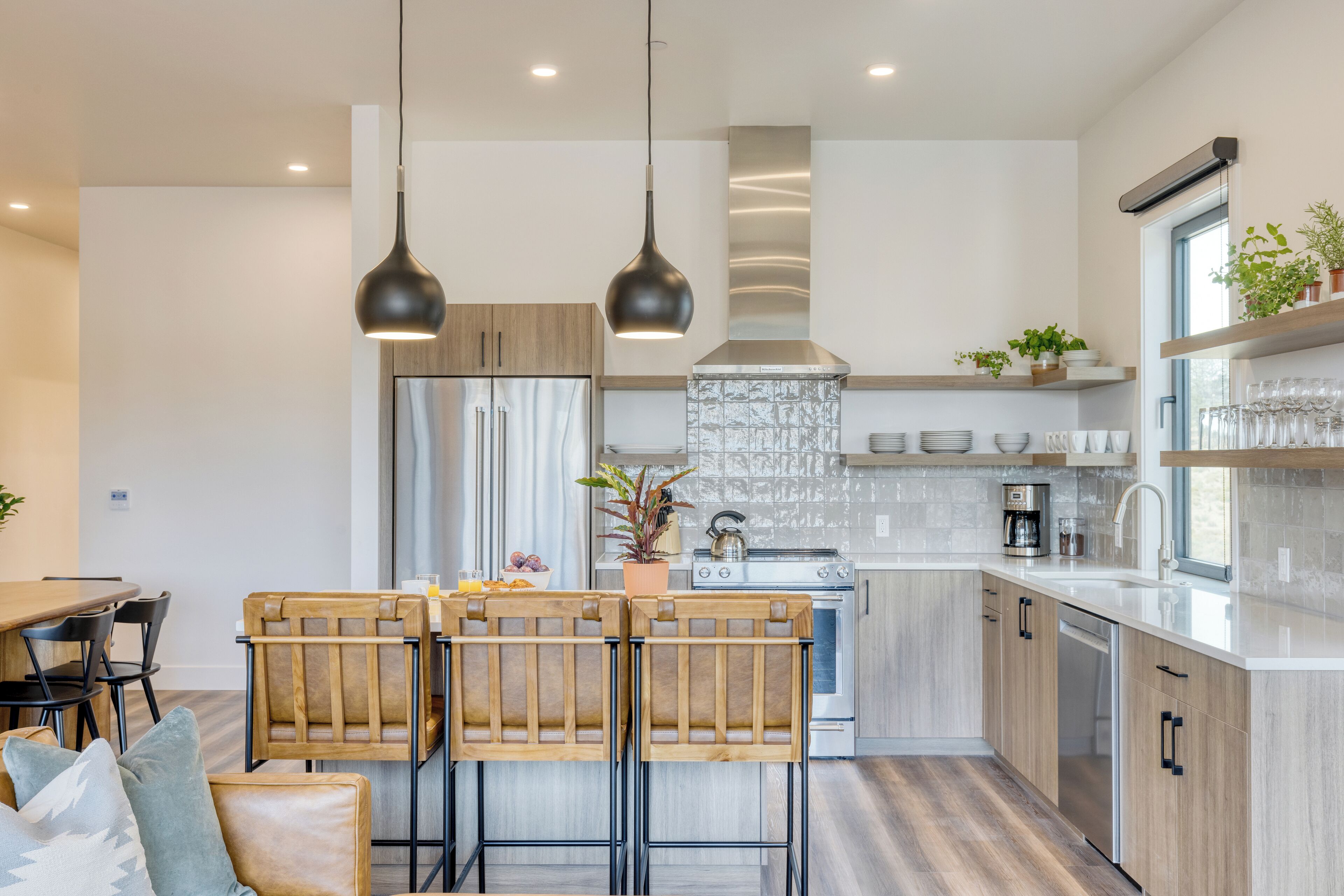 A modern kitchen with wooden cabinets, a stainless steel refrigerator, a gas stove, and a built-in oven. There are two bar stools, a dining table, and a potted plant.