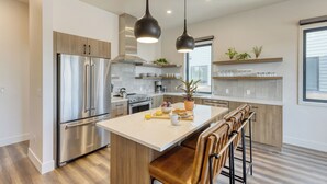 A modern kitchen with stainless steel appliances, a central island with bar stools, pendant lighting, and a window with blinds.