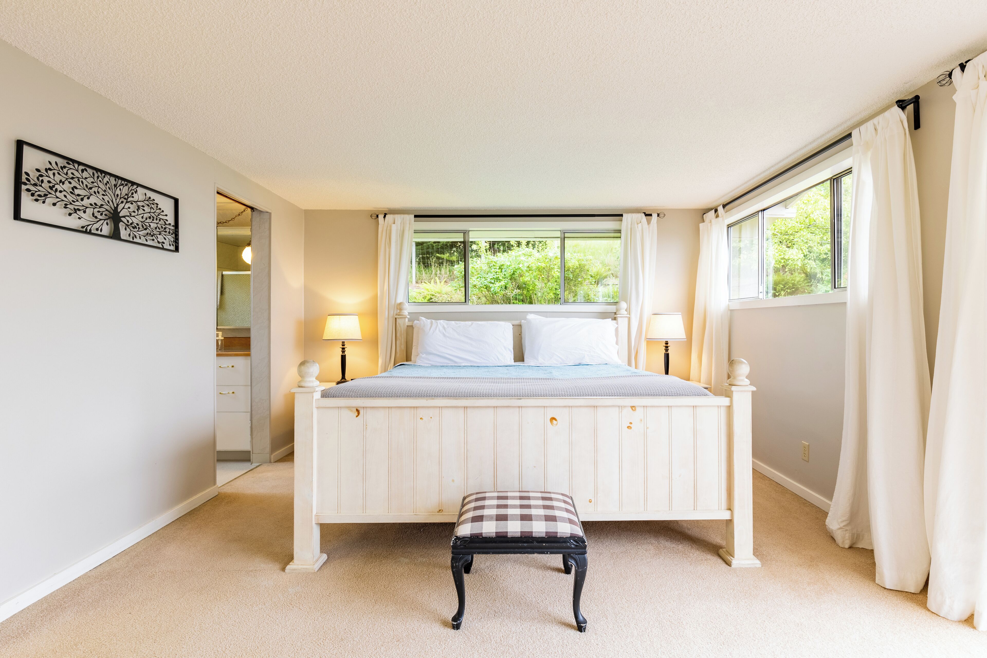 A bedroom with a bed, bedside lamps, a bench, a window with curtains, and a wall-mounted artwork.