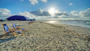 A beach with blue lounge chairs and a pier in the background. - Aloha Ohana by AvantStay | Private Pool Near Beach (Port Aransas)