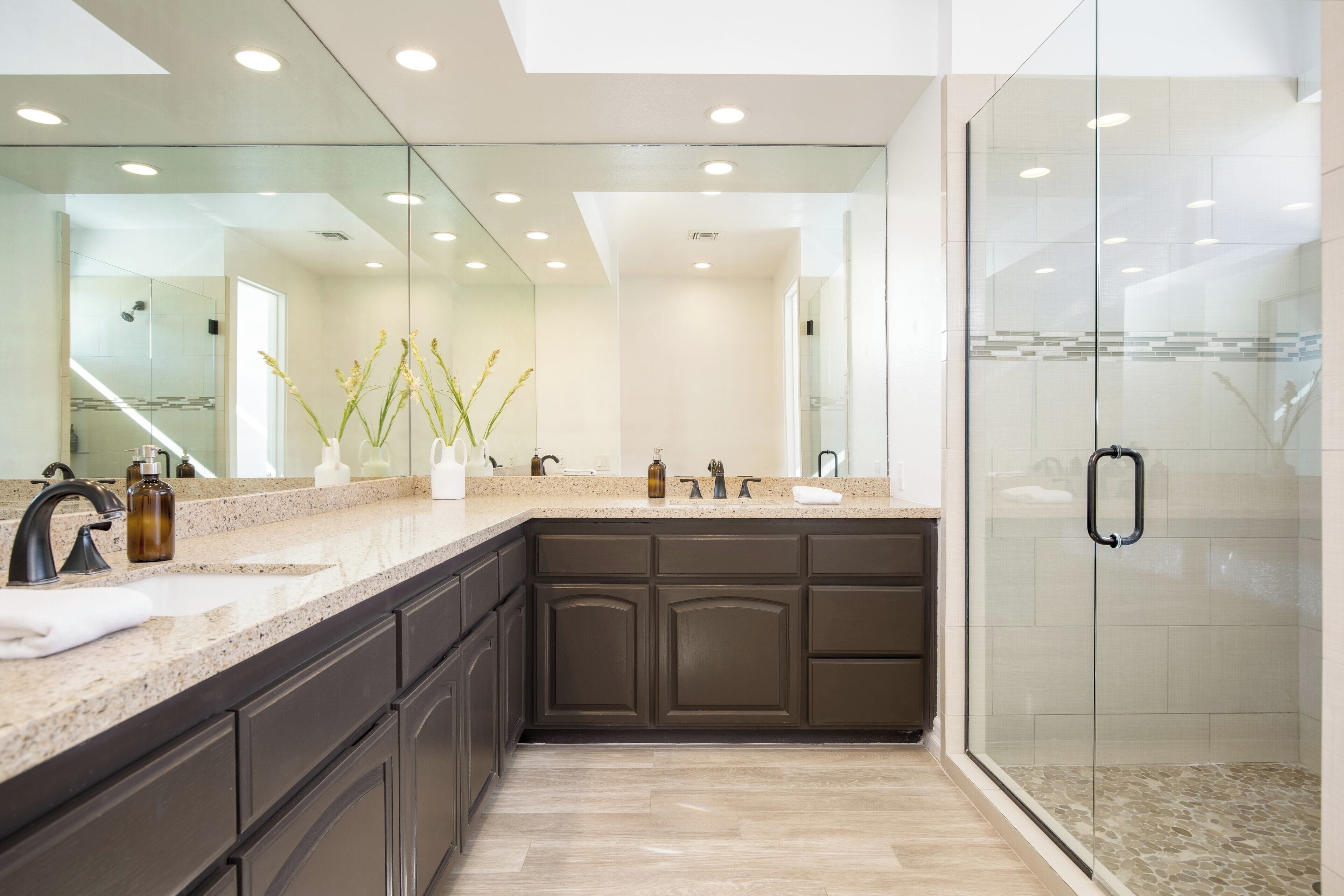 A modern bathroom with a large mirror, dark cabinets, a glass shower enclosure, and a granite countertop.