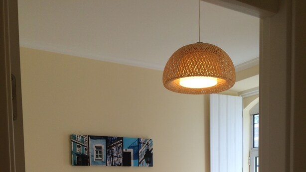 Een net opgemaakt bed met witte lakens en twee kussens, een geweven hanglamp erboven en een lijst met een stadsgezicht aan de muur.