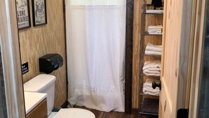 A compact bathroom with a toilet, a shelf with towels, and a shower curtain. - Dinosaur Valley RV Park Cabin 3 (Glen Rose)
