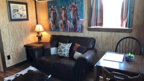 A cozy living room with a leather couch, a small table, a lamp, a painting of horses, and a window with curtains. - Dinosaur Valley RV Park Cabin 3 (Glen Rose)