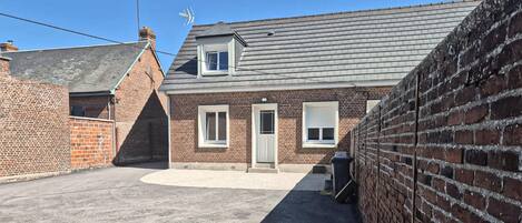 Une maison à deux étages, construite en briques, avec un toit gris, une porte d’entrée et un petit jardin devant.