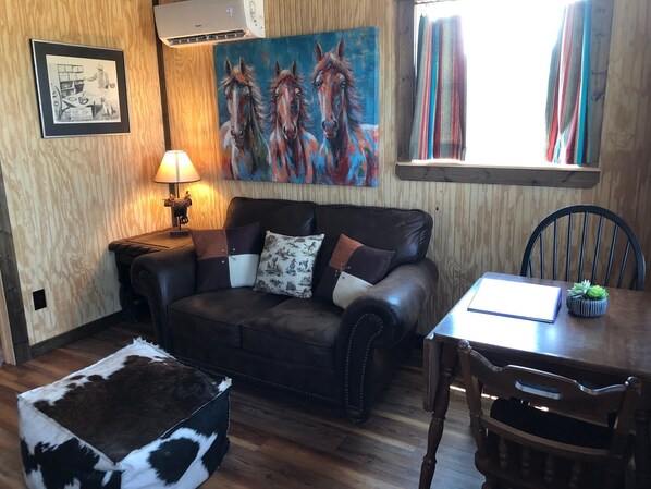 Una sala de estar acogedora con un sofá de cuero, una mesita, una lámpara, un cuadro de caballos y una ventana con cortinas.
