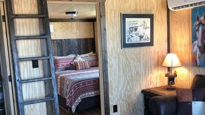 A rustic room with a wooden ladder, a bed with patterned bedding, a cowhide ottoman, a leather sofa, and a lamp on a side table. - Dinosaur Valley RV Park Cabin  4 (Glen Rose)
