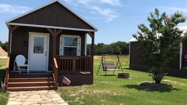 A small wooden cabin with a porch, a flag, and a swing set in a grassy area. - Dinosaur Valley RV Park Cabin  4 (Glen Rose)