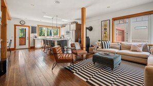 A spacious living area with a kitchen, wooden flooring, a striped rug, and a large window with curtains.