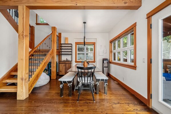 A spacious room with a wooden staircase, a dining area with a table and chairs, and large windows providing natural light.