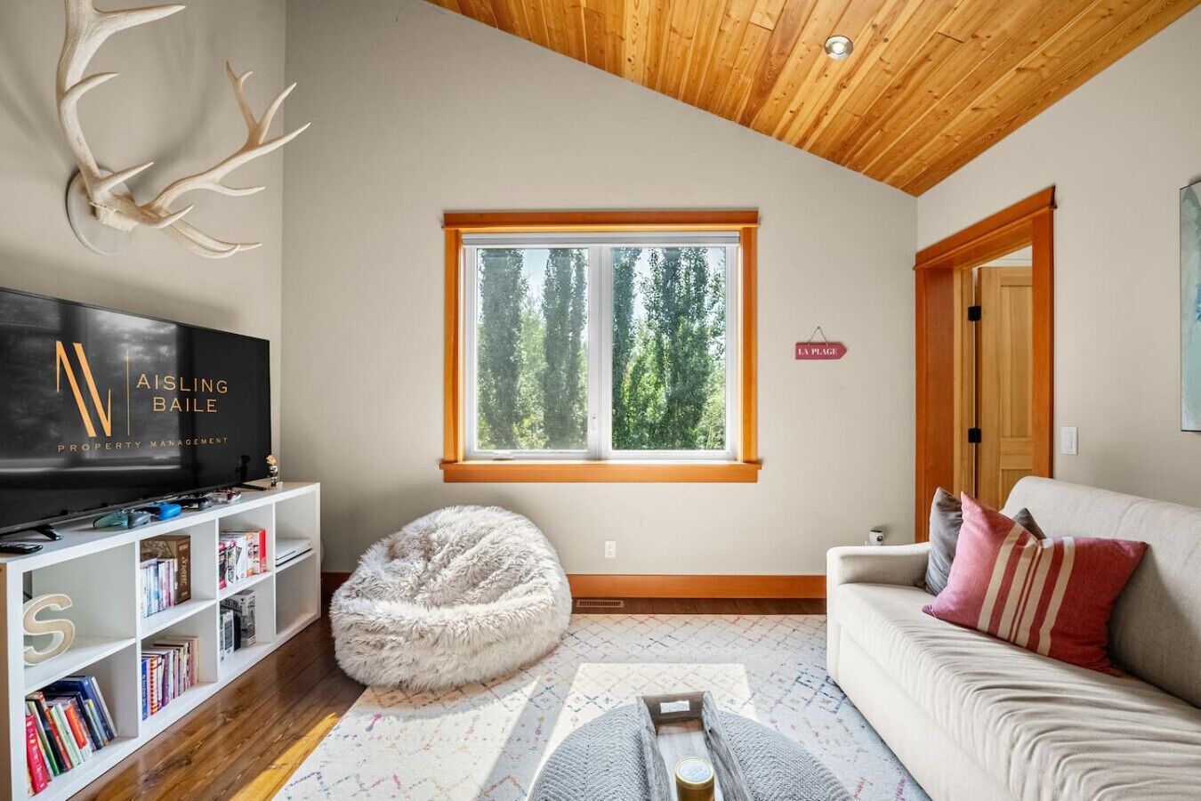 Una sala de estar moderna con una televisión, estanterías, un sillón de peluche, un sofá con cojines, un ventanal grande y un techo de madera.