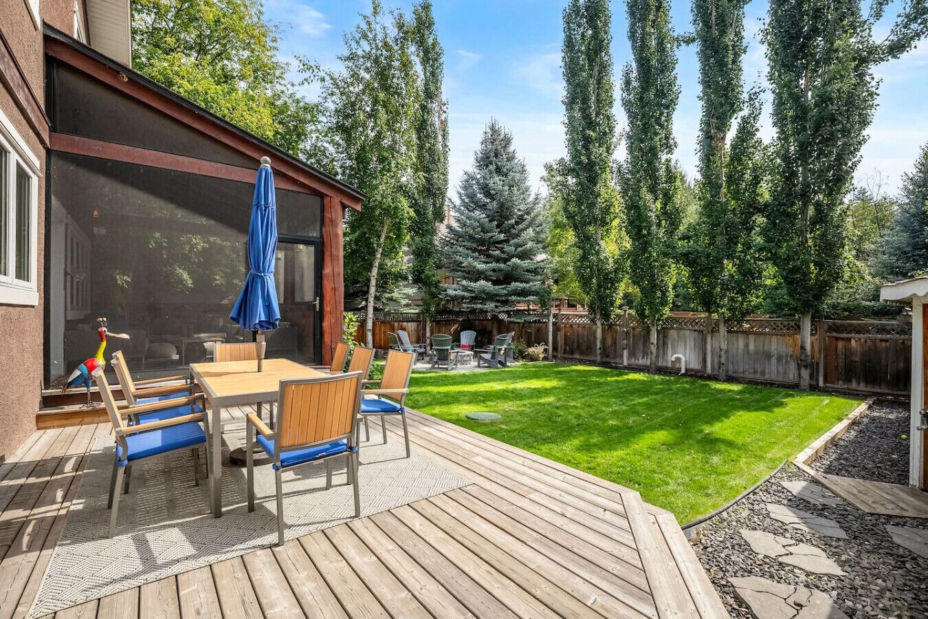 A backyard with a wooden deck, a dining table with chairs, a blue umbrella, and a bird sculpture.