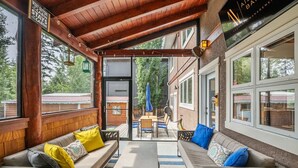 A covered outdoor seating area with wooden benches and a table, large windows, and a view of the surrounding area.