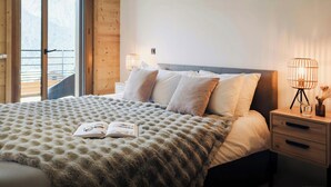 A bedroom with a large bed, a nightstand, two bedside lamps, and a window with a view of mountains. - Loft Petit Charbel - Charming stay with balconies - OVO Network (Châtel)