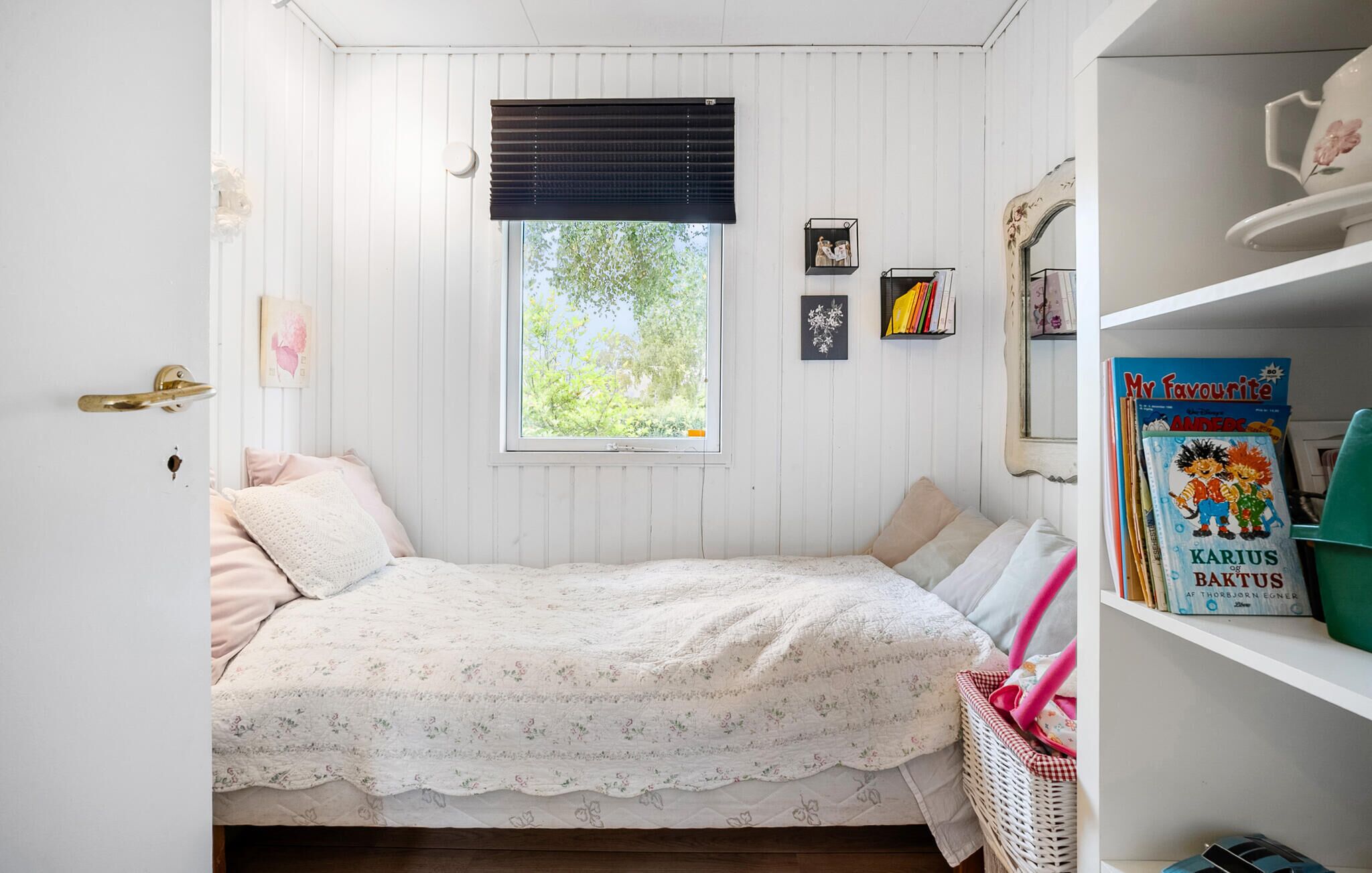 Une chambre confortable avec un lit simple, une fenêtre avec des stores, une bibliothèque remplie de livres et une commode avec un miroir.