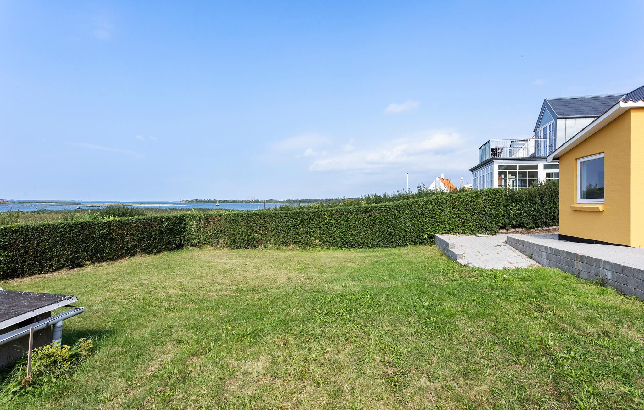 A grassy backyard with a hedge, a yellow building, and a view of the sea.