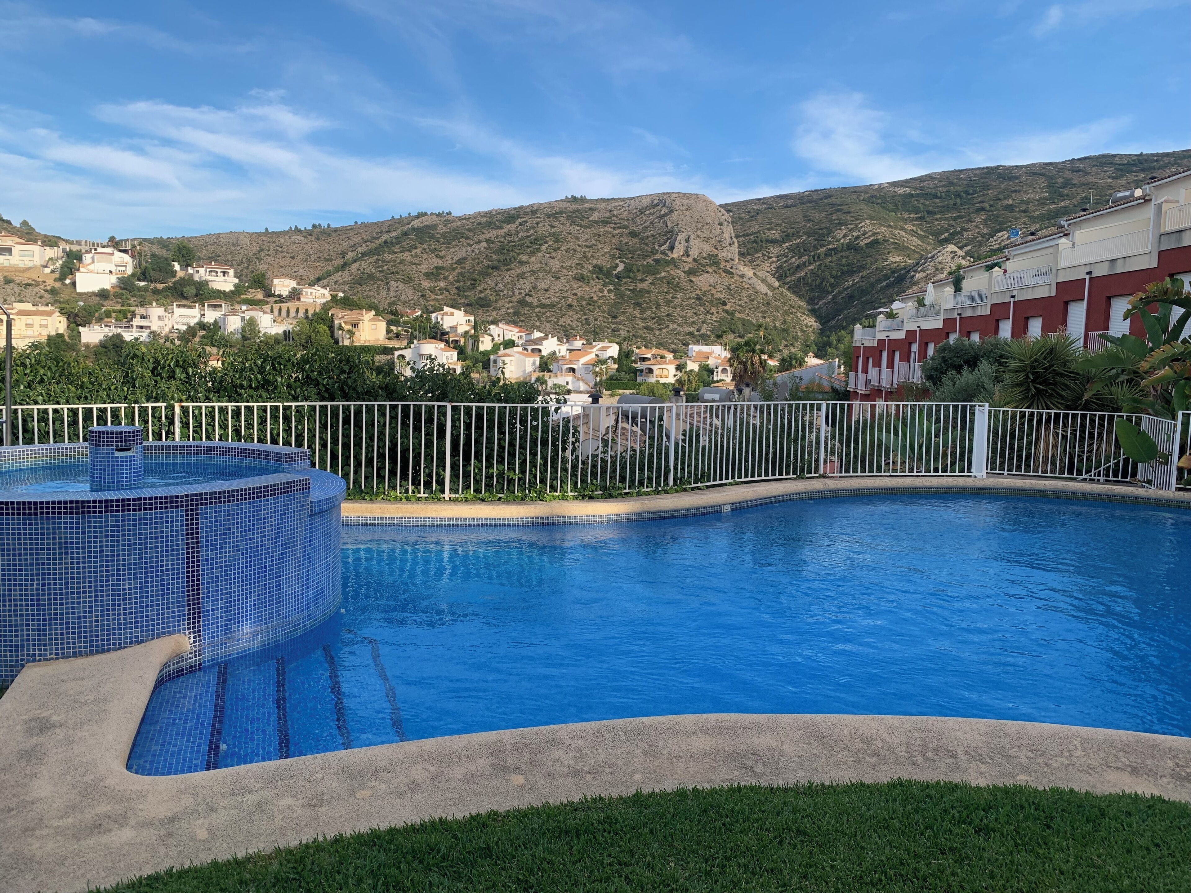 A swimming pool with a tiled hot tub, surrounded by a white fence, with a view of a hillside town in the background.