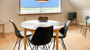 Un coin repas avec une table ronde, des chaises noires, un luminaire suspendu rouge, une fenêtre donnant sur un espace verdoyant et un téléviseur posé sur une petite table.
