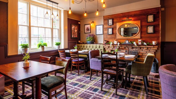 A dining area with a wooden table and chairs, a patterned rug, and a brick wall with a mirror and framed pictures. - The Bell Hotel  (Burgh le Marsh)