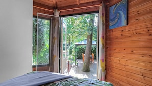 Une pièce avec un lit, des murs en bois, une fenêtre avec des rideaux et une vue sur un espace extérieur comprenant un arbre et une table.