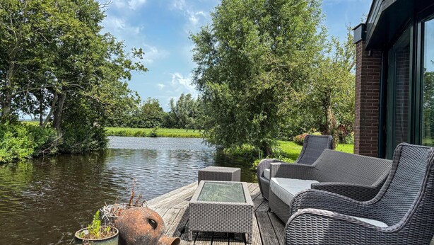 A wooden deck with wicker furniture overlooking a river, surrounded by lush greenery. - Amazing home in Stolwijk with house sea view (Stolwijk)