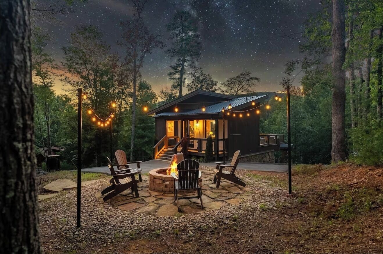 A cabin with a fire pit and outdoor seating, surrounded by trees and string lights.