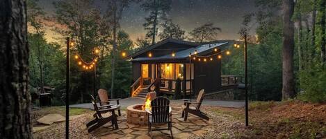 A cabin with a fire pit and outdoor seating, surrounded by trees and string lights.