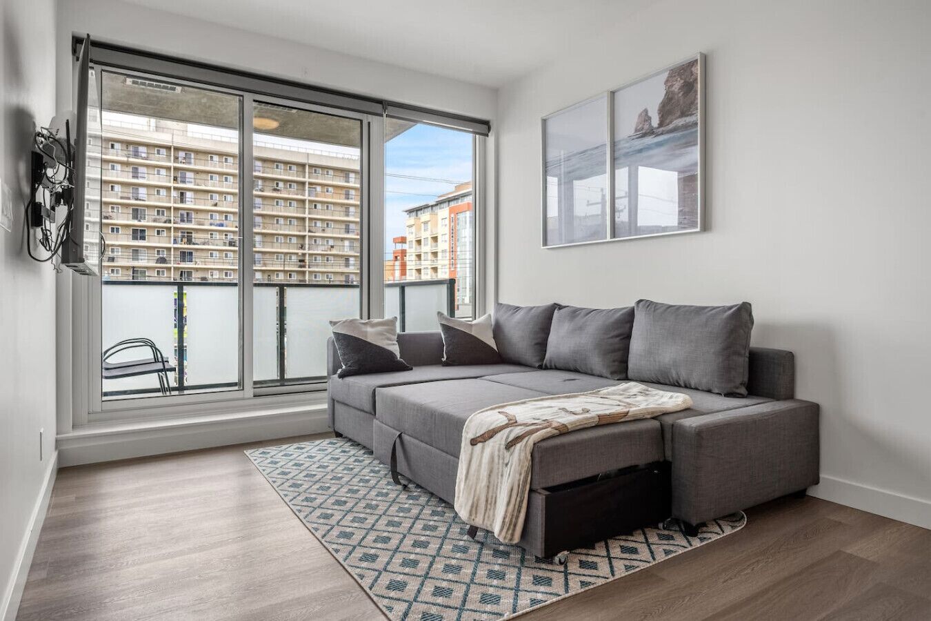 A modern living room with a grey sectional sofa, a patterned rug, and a large window offering a view of a high-rise building.