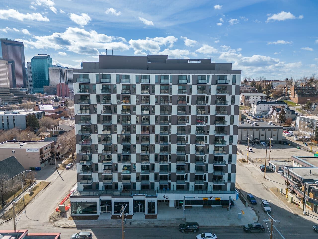 A high-rise building with a grid of balconies, surrounded by other buildings and a parking lot.