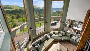 A room with a large sectional sofa, multiple windows offering views of a green landscape, and a deck area visible through one window. - The Deadwood Lookout-Sleeps 22 (Sturgis)