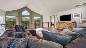 A spacious living room with a high ceiling, large windows offering a view of rolling hills, a wooden entertainment unit, and a comfortable seating area. - The Deadwood Lookout-Sleeps 22 (Sturgis)