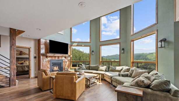 A spacious living room with a fireplace, large windows, and a view of the outdoors. - The Deadwood Lookout-Sleeps 22 (Sturgis)