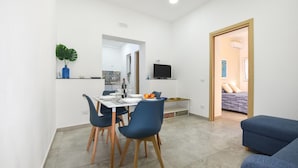 Un espace de vie compact comprenant un coin repas, un téléviseur fixé au mur et une chambre à coucher visible à travers une porte ouverte.