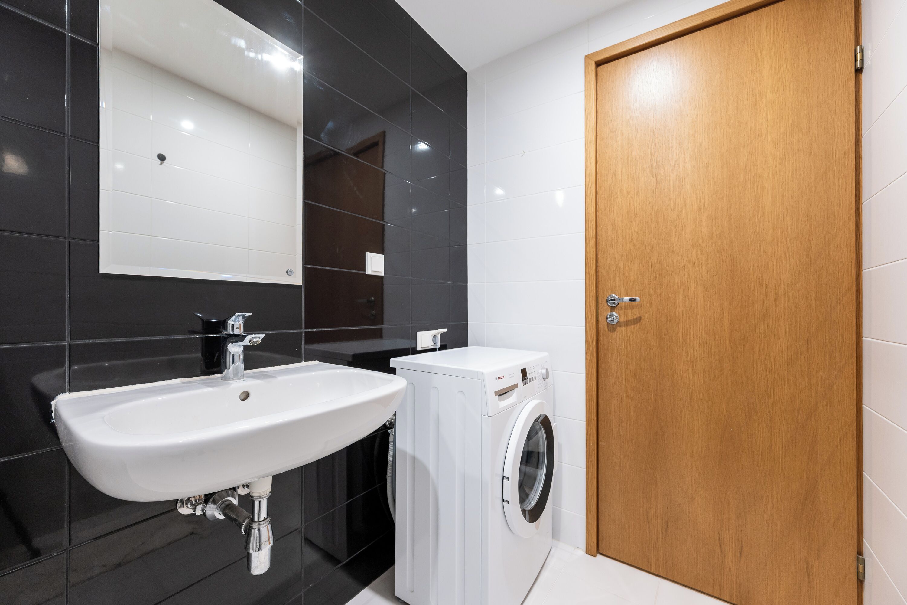 A modern bathroom with a white sink, a washing machine, and a wooden door.