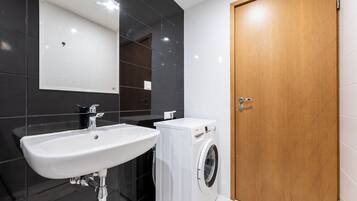 A modern bathroom with a white sink, a washing machine, and a wooden door.