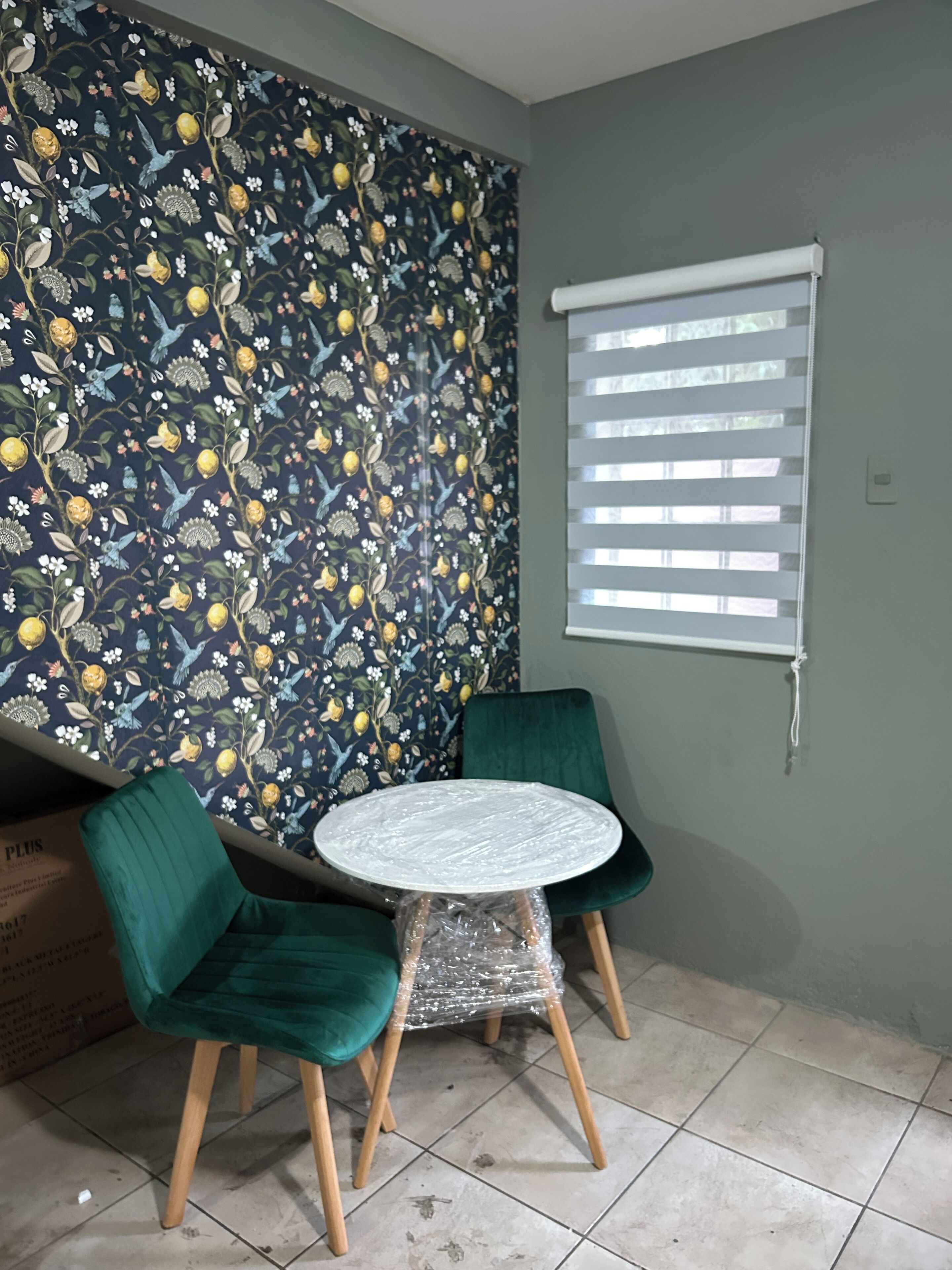 Une pièce avec un papier peint à motifs floraux, une table ronde entourée de chaises, une fenêtre avec des stores et un fauteuil vert.