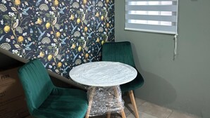 A room with a floral wallpaper, a round table with chairs, a window with blinds, and a green chair.